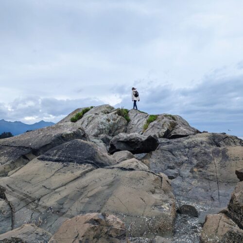 Gabi, the Fringe Explorer, walking on top of a rock feature on a beach. The sky is dreary and cloudy