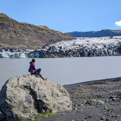 Gabi, The Fringe Explorer, sitting on a rock in front of a glacier. It's a sunny clear day