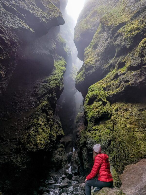 Woman wearing a grey beanie and red puffer jacket is sitting on a rock looking into a foggy moss covered gorge