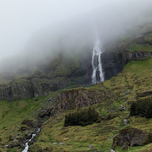 Waterfall pouring down a mountainside partially hidden by foggy clouds. The foreground is uneven moss and grass-covered rock.